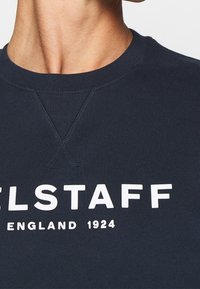 Gros plan d’un t-shirt bleu marine à col rond avec un texte blanc indiquant "BELSTAFF ENGLAND 1924" sur la poitrine.