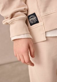 Lapsen käsi rentona beige raidallisessa hihassa, jossa on musta "Superdry"-logopatchi hihassa.