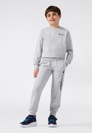Grijze set van een sweatshirt en een joggingbroek met elastische tailleband, met een donkerblauw logo op de broek. Draagt daarbij marineblauwe sneakers.