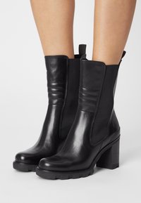 Bottines en cuir noir avec des panneaux élastiques sur les côtés, des accents texturés et un talon épais. Fini lisse avec une semelle en caoutchouc à rainures.