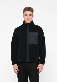 Black fleece jacket with a high collar and zipper, featuring a matte black chest pocket and contrasting textures. Soft, warm material.