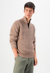 Beiger Strickpullover mit einem halben Reißverschluss, gerippter Textur und langen Ärmeln. Getragen über einem dunklen Oberteil und kombiniert mit grünen Hosen.