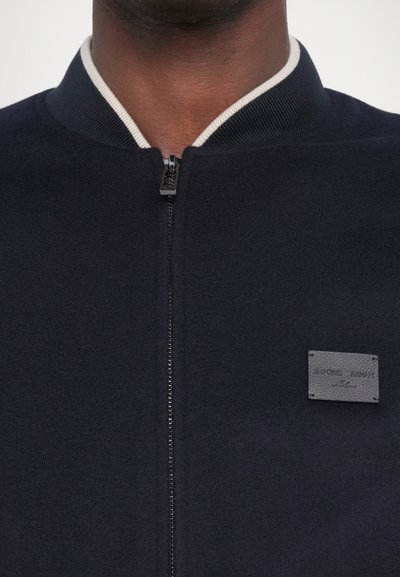 Tume sinine pommitaja jakk musta tõmblukuga, helehall ribidega krae ja tekstuuriga kinnitus "Emporio Armani" vasakul rinnal.
