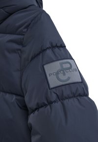 Marinblå puffjacka med ärmar som har quiltade sektioner och en grå tygpatch med präglad "POLO CLUB"-logotyp. Mjuk och slät textur.