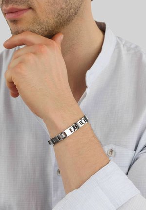 Joven con camisa blanca apoyando la barbilla en su mano, con una pulsera de eslabones metálicos plateados en la muñeca.