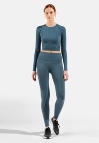 Langärmliges, gekürztes Trainingsoberteil und passende, hochgeschnittene Leggings aus einem sanften, gerippten Stoff, beide in einer gedämpften Tealfarbe. Schwarze Sportschuhe.