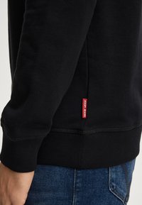 Felpa nera, tessuto morbido, polsini e orlo a coste, etichetta laterale con "JOOP! JEANS" in rosso, design aderente, texture liscia.