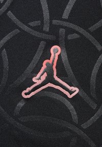 Una tela negra texturizada presenta un patrón abstracto en gris. Un logo Jumpman en color rosa se muestra de manera prominente en el centro, destacando los elementos de diseño atlético.