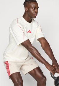 Jasny sportowy t-shirt z czerwonymi akcentami i logo, w połączeniu z beżowymi szortami z czerwonymi paskami, noszony podczas ćwiczeń.