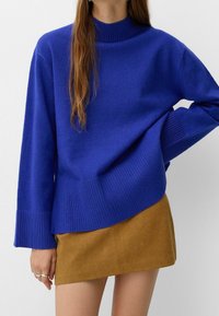 Bright blue knit sweater with a high collar, ribbed cuffs, and an asymmetrical hem, paired with a tan suede mini skirt.