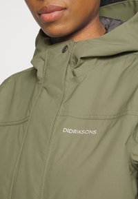 Gros plan d'une personne portant une veste Didriksons vert olive avec un col montant et une capuche.