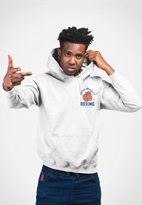 Grå hoodie med en framficka, prydd med en boxningsgrafik och textdesign i blått och orange. Bärs av en person med kort hår.
