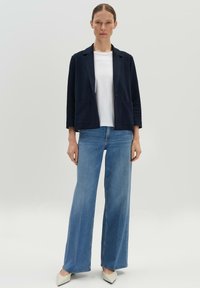 Navy Blazer mit zwei Vordertaschen, weißem T-Shirt und weit geschnittenen hellblauen Jeans. Ein Paar spitz zulaufende weiße Schuhe vervollständigt das Outfit.