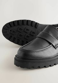 Mocasines de cuero negro con un acabado texturizado, que presentan una suela de goma gruesa con un agarre patentado y un diseño de punta redondeada y elegante.