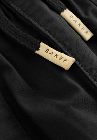 Nahaufnahme von schwarzem Stoff mit zwei schwarzen Zugbändern, die jeweils mit beigen Endstücken versehen sind, auf denen "BAKER" in schwarzer Schrift steht.