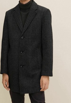 Homme portant un manteau texturé gris foncé boutonné par-dessus un pull à col roulé noir, sur un fond clair.