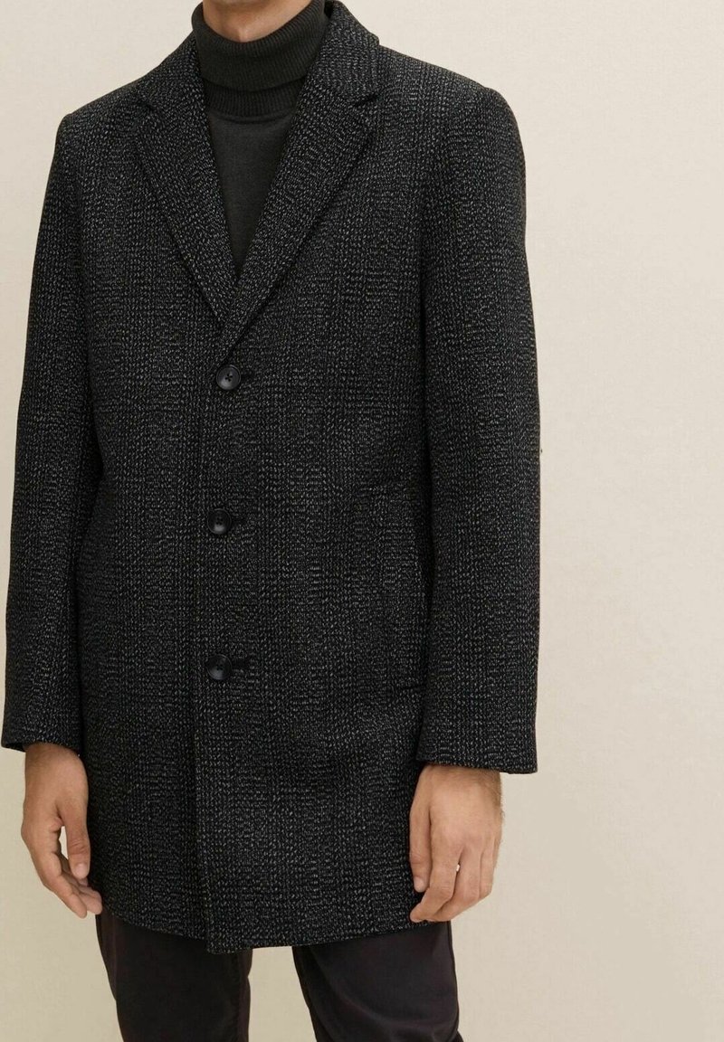 Homme portant un manteau texturé gris foncé boutonné par-dessus un pull à col roulé noir, sur un fond clair.