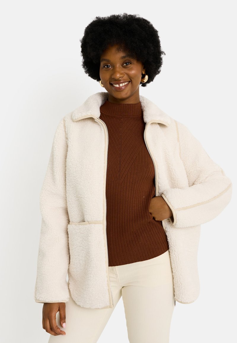 Chaqueta de forro polar color crema con cremallera al frente, dos bolsillos delanteros y una textura suave. Cuenta con un cuello y piping beige en contraste.