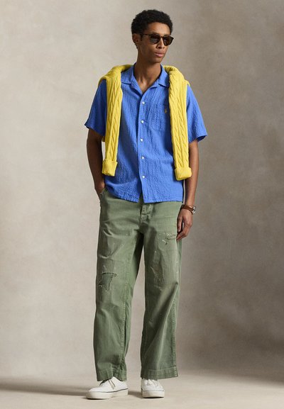Homme portant des lunettes de soleil, une chemise bleue à manches courtes, un pull jaune drapé sur les épaules, un pantalon vert déchiré et des baskets blanches, debout sur un fond neutre.