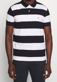 Polo shirt rayé noir et blanc, manches courtes, col avec fermeture à boutons, présentant un petit logo sur la poitrine gauche. Tissu en coton.
