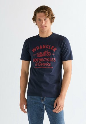 Młody mężczyzna o kręconych włosach nosi granatowy T-shirt z czerwonym napisem "Wrangler Motorcycles & Service" oraz grafiką motocykla, w połączeniu z niebieskimi dżinsami.