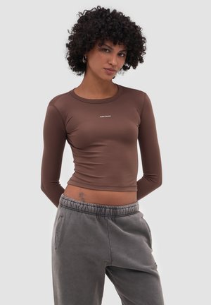 Mujer con cabello rizado, vestida con un top corto ajustado de manga larga marrón y pantalones deportivos grises sueltos, de pie frente a un fondo liso.