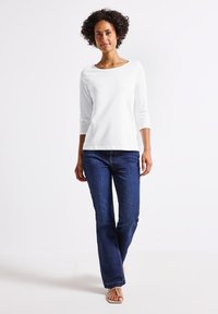 Witte blouse met lange mouwen en een bootnek, gecombineerd met donkerblauwe denim jeans. De jeans hebben een klassieke snit en zichtbare stiksels.