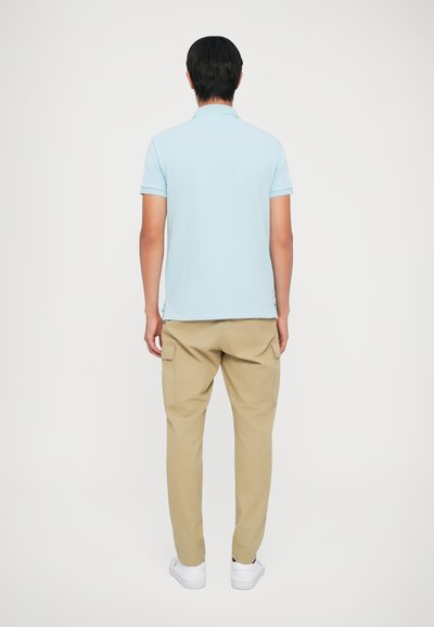 Άνδρας με ανοιχτό μπλε κοντομάνικο πόλο, μπεζ cargo παντελόνι και λευκά αθλητικά παπούτσια, όρθιος μπροστά σε ένα λευκό φόντο.