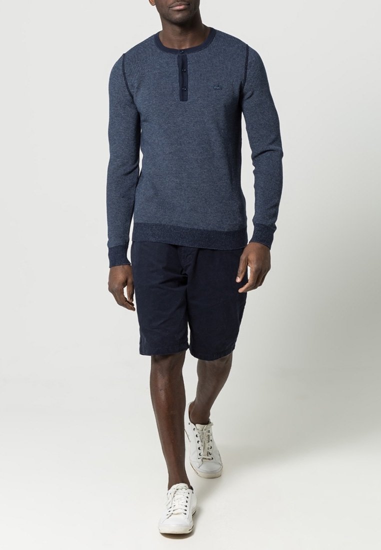 Homme marchant portant un t-shirt henley bleu à manches longues, un short bleu marine et des baskets blanches sur un fond clair uni.
