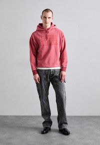 Czerwona bluza z kapturem z nadrukiem "J'ADORE ARIES", połączona z luźnymi szarymi dżinsami. Model nosi czarne buty i stoi na tle neutralnej ściany.