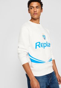 Hvid sweatshirt med et blåt "Replay" logo og et holdlogo, prydet med blå bølgeaccentuer. Blødt stof med ribbede manchetter og kant.