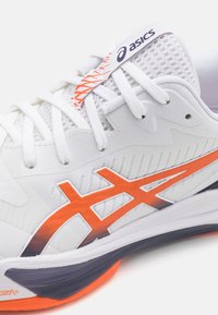 Hvit atletisk sneaker med teksturert overdelen, oransje aksentlogo og en polstret såle med en mørk lilla bunn.