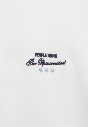 Bílá látka s černým vyšitým nápisem „Lidé si myslí, že mám silné názory“ nad třemi světle modrými hvězdami.