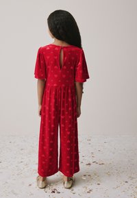 Tuta rossa con maniche corte a volant e schiena con taglio a goccia, caratterizzata da un motivo a cuori su tutto il tessuto morbido e vellutato; pantaloni a gamba larga.