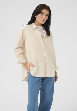 Light beige shirt with a button-up front, long sleeves, and a curved hem. Worn over a white top and paired with blue jeans.