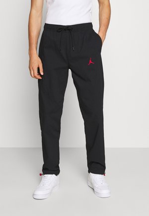 Person iført sorte snøre bukser med rødt logo på venstre lår og hvide sneakers, stående mod en ensfarvet baggrund.