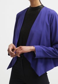 Lila, löst sittande kavaj med V-ringning och vida ärmar, bärs över en svart, texturerad topp. Händerna vilar på höfterna.