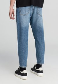 Jeans in denim blu con una vestibilità comoda, texture sbiadita e dettagli strappati sul retro. Indossati con sneakers nere e bianche.