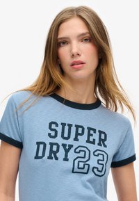 Lichtblauw T-shirt met korte mouwen en een zwarte geribde kraag en manchetten. Voorkant met opvallende zwarte tekst: "SUPER DRY 23." Zachte stoftextuur.