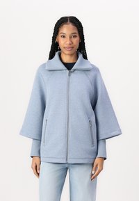 Lyseblå zip-up jakke med høy krage, trekvart ermer, to sidelommer og en myk tekstur.