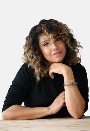 Haut à manches longues côtelé noir ; cheveux châtains ondulés avec des reflets blonds ; portant plusieurs bracelets avec des perles et des accents métalliques au poignet.