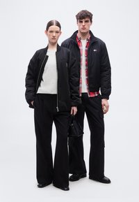 Δύο μοντέλα στέκονται το ένα δίπλα στο άλλο, φορώντας μαύρα μπουφάν τύπου bomber και μαύρο παντελόνι· η γυναίκα κρατάει μια μαύρη τσάντα, ενώ ο άντρας φοράει κόκκινο πουκάμισο καρό κάτω από το μπουφάν.