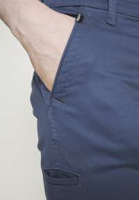 Ciemnoniebieskie spodnie z gładkiej, teksturowanej tkaniny. Posiadają boczną kieszeń z widocznym przeszyciem oraz małą metkę marki przy otworze kieszeni.