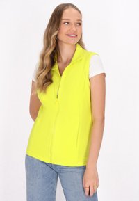 Neon-gelber ärmelloser Vest mit hohem Kragen, Frontreißverschluss und glatter Textur. Kombiniert mit einem weißen kurzärmeligen Hemd und Jeans.