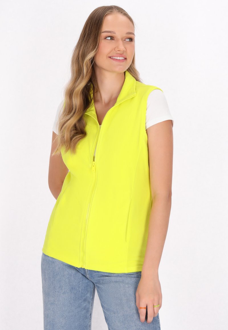 Neon-gelber ärmelloser Vest mit hohem Kragen, Frontreißverschluss und glatter Textur. Kombiniert mit einem weißen kurzärmeligen Hemd und Jeans.
