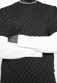 Černé tričko s krátkým rukávem s texturovaným vzorem, položené na bílém tričku s dlouhým rukávem. Má kulatý výstřih a štítek s logem.