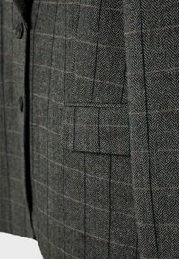 Blazer gris à motif chevron avec de subtils carreaux ; il dispose d'une poche poitrine unique et de boutons noirs à l'avant. La texture semble tissée.