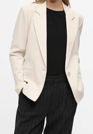 Femme portant un blazer crème sur un haut noir et un pantalon sombre à fines rayures, les mains dans les poches.
