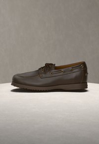 Chaussure bateau en cuir brun foncé avec un détail lacé et une semelle en caoutchouc sur une surface claire avec un fond neutre.