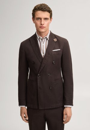 Junger Mann, der einen dunklen, zweireihigen Blazer mit einem hell gestreiften Hemd trägt, steht vor einem einfarbigen hellen Hintergrund.
